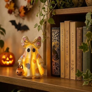 May include: A cream-colored ghost figurine with a large bow on its head, decorated with pink floral patterns and colorful patches. The ghost is illuminated from within and holds a small orange jack-o'-lantern bucket in its hand.