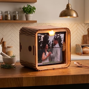 Op de afbeelding: Een lichtgekleurde houten dooslijst met een ingebouwd lampje aan de bovenkant, met een foto van een koppel in een bruiloftssetting. De lijst heeft een tandwielmechanisme aan de zijkant en een glad, afgerond ontwerp met decoratieve uitsparingen.