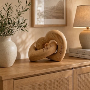 Op de afbeelding: Een decoratief houten beeldhouwwerk bestaande uit twee in elkaar grijpende hartvormen, gesneden uit lichtgekleurd hout met een zichtbare nerf. De namen Olivia en Liam zijn in cursief schrift gegraveerd op het oppervlak van de houten harten.