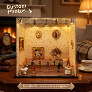 May include: A miniature museum display case featuring illuminated spotlights, gold-toned ornate picture frames on patterned wallpaper, and small figurines standing behind a red velvet rope barrier. The display includes white pedestals with floral arrangements.