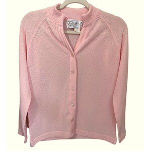 May include: A light pink long-sleeve cardigan with a ribbed mock neck collar and a button-down front. The garment features a textured knit fabric and a white interior label with the text Talbott Traveler. The design includes raglan sleeves.