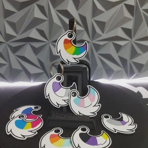Può includere: Una collezione di sette portachiavi a forma di mezzaluna con vari motivi colorati. Ogni portachiavi presenta un bordo bianco e un anello di metallo fissato sulla parte superiore. I design includono strisce arcobaleno, pastello e multicolori.