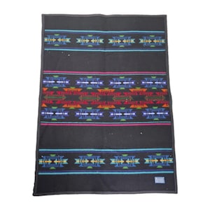 May include: A rectangular black blanket featuring horizontal bands of geometric patterns in shades of red, orange, green, blue, and yellow. The design includes thin turquoise and magenta stripes, with a small rectangular label in the bottom corner.