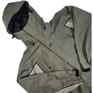 May include: An olive green Helly Hansen hooded jacket featuring a black interior lining, a front zipper closure with a protective storm flap, adjustable drawstrings on the hood, and contrasting beige panels on the sleeves and pockets.