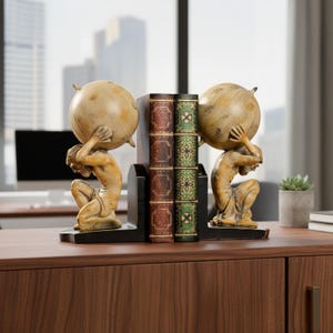 Puede incluir: Un par de sujetalibros con dos figuras idénticas en posición de rodillas, cada una sosteniendo un gran globo terráqueo beige desgastado sobre sus hombros. Las figuras se asientan sobre bases negras, flanqueando dos libros decorativos con lomos marrón y verde.