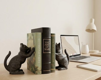 May include: Two dark gray cat-shaped bookends made of cast metal, positioned on either side of three books titled Cotswold Tales, The Art of War, and The Secret Garden, with textured fur details and arched tails facing outward on a desk.