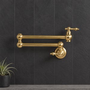Op de afbeelding: Een aan de muur gemonteerde pannenvuller-kraan met een geborstelde messing afwerking, voorzien van een dubbel scharnierende arm, een ronde basisplaat en twee hendels voor het regelen van de waterstroom en temperatuur in de keuken.