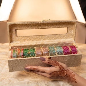 May include: A rectangular gold glitter storage box with a clear rectangular window on the lid, containing a row of multicolored glass bangles. The bangles feature thin gold wire accents and small gold beads arranged in a repeating pattern.