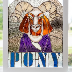 May include: A stained glass panel in a white frame featuring a goat with large curved horns, orange ears, and a purple chest. The background is textured clear glass. The word PONY is written in bold blue letters at the bottom of the panel.