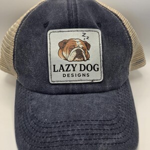 May include: A charcoal grey baseball cap with a tan mesh back and a rectangular patch on the front. The patch features a brown and white bulldog sleeping with Zzz symbols above it and the text LAZY DOG DESIGNS printed in black lettering.