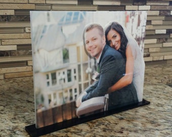 May include: A black acrylic stand with a photo of a couple. The couple is standing in front of a building with a blurred background. The woman is wearing a white dress and the man is wearing a suit.