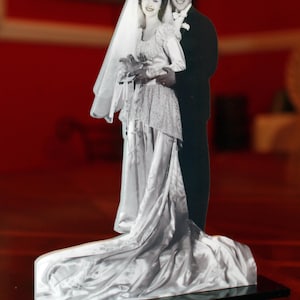 May include: A black and white wedding photo of a couple standing together. The bride is wearing a white dress and veil, and the groom is wearing a suit. The photo is mounted on a black base.