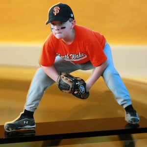 May include: A young baseball player wearing a black and orange baseball cap with the letters "SF" on it, a red and orange shirt with the words "North Wake" on it, grey pants, and a black and brown baseball glove. The player is in a fielding position with one leg bent and the other leg straight.
