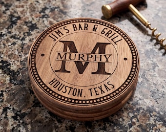 May include: A round wooden coaster featuring a laser-engraved design with a large letter M, the name MURPHY, and the text JIM'S BAR & GRILL HOUSTON, TEXAS. The coaster has a decorative dotted border and a smooth, natural wood grain finish.
