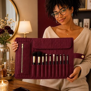 May include: A deep burgundy quilted makeup brush roll featuring a vertical zippered pocket with gold hardware and multiple individual slots containing various cosmetic brushes with black handles and gold ferrules, secured by a side snap closure.