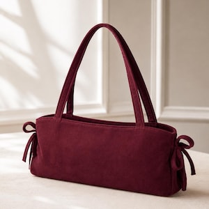 May include: A deep burgundy rectangular handbag with a soft, matte texture. The bag features two upright shoulder straps and decorative fabric ties on both the left and right sides. The design is structured with a flat base and clean, simple lines.