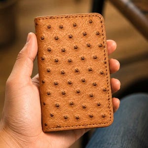 May include: A rectangular tan wallet featuring a distinct textured surface with evenly spaced raised quill follicles. The wallet has rounded corners and visible stitching along the edges, showcasing a classic bifold design in a warm brown tone.