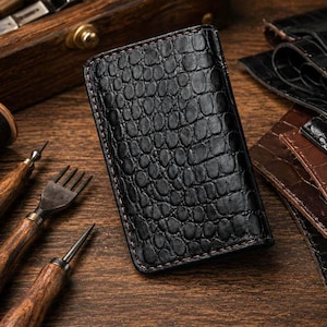 May include: A rectangular black leather card holder featuring a textured crocodile skin pattern. The accessory is constructed with visible dark stitching along the edges, showcasing a structured design with a glossy finish on the exterior surface.