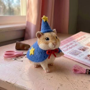 May include: A needle-felted hamster figurine wearing a blue wizard hat with a yellow star, a matching blue cape with a yellow star, and a red bow tie. The hamster has light brown and white fur, black eyes, and a small pink nose.