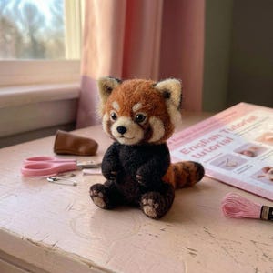 Könnte beinhalten: Eine nadelgefilzte rote Panda-Figur mit rostfarbenem Kopf, weißen Gesichtsmarkierungen und einem dunkelbraunen Körper. Das kleine Tier sitzt in aufrechter Position mit sichtbaren Pfotenballen und hat eine strukturierte Wolloberfläche.