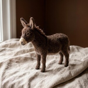 May include: A needle-felted donkey figurine crafted from dark brown wool fibers. The animal figure features long, upright ears, a short mane, and a lighter tan-colored muzzle. The texture of the wool is visible throughout the body.