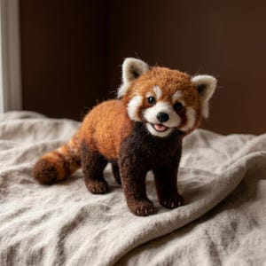May include: A needle-felted red panda figurine with a textured, fuzzy coat in shades of reddish-brown and dark brown. The creature features white markings around its eyes and muzzle, pointed ears, and a small, open mouth showing a pink tongue.