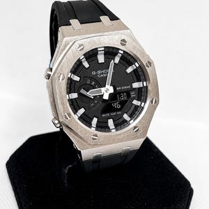 May include: A G-Shock Casio watch with a silver-toned octagonal metal bezel featuring eight visible screws. The watch has a black dial with white hour markers, silver hands, and a digital display window. It includes a black resin strap.