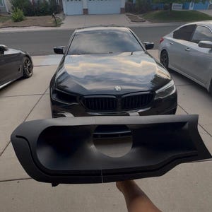 May include: A matte black plastic automotive bumper air intake grille insert featuring a central circular opening, held in front of a black BMW sedan parked on a concrete driveway in a residential neighborhood setting.