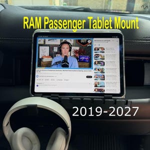 May include: A black RAM passenger tablet mount for 2019-2027 vehicles, featuring a tablet displaying a video interface. A pair of white over-ear headphones rests in the foreground. The text RAM Passenger Tablet Mount 2019-2027 is visible.
