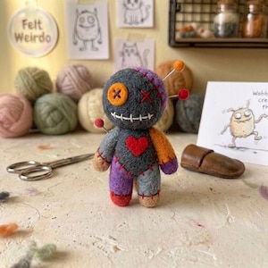 Könnte beinhalten: Eine nadelgefilzte Puppe mit dunkelgrauem Körper, einem leuchtend orangefarbenen Knopfauge, einem roten gestickten Auge und einem weißen gestickten Mund. Sie hat ein rotes Herz auf der Brust, violette und graue Gliedmaßen und bunte Nadeln am Kopf.