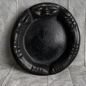 May include: A circular black wooden tray featuring a carved face at the top centre and a series of repeating geometric spike-like patterns etched along the raised outer rim. The surface of the tray is smooth and dark with a visible wood grain texture.