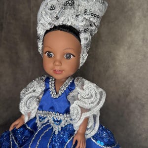 May include: A doll with dark skin and black hair wearing a tall, silver sequined headpiece. The doll is dressed in a royal blue gown featuring silver lace trim, sheer ruffled sleeves, and intricate sequin embellishments on the bodice and skirt.