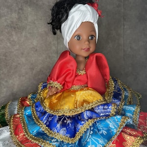 May include: A doll with dark curly hair wearing a white headwrap and a multi-layered dress. The dress features a red bodice with puffed sleeves, a yellow middle tier, and blue and red bottom tiers, all accented with gold metallic trim.