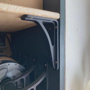 May include: A matte black L-shaped shelf bracket mounted to a dark vertical surface, supporting a light-colored wooden shelf. The bracket features a reinforced triangular design with visible screw holes and metallic hardware fasteners.