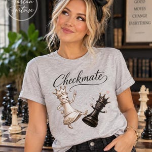 May include: A light gray short-sleeve t-shirt featuring a graphic of a white chess queen piece kicking a black chess queen piece. The text "Checkmate" is printed in black cursive script above the illustrated chess pieces on the front.