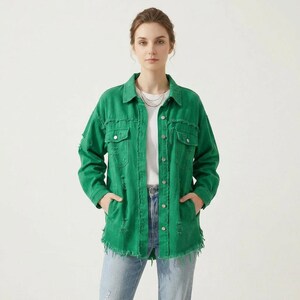 Pode incluir: Um casaco de ganga verde brilhante com gola, mangas compridas e fecho frontal de botões. O casaco inclui dois bolsos no peito com abas abotoadas, detalhes desgastados e uma bainha desfiada, usado sobre uma camisa branca lisa.