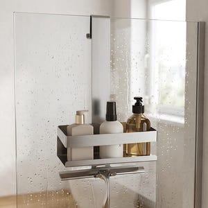 May include: A silver metal shower caddy hangs over a glass shower door. The rectangular shelf holds three bottles: a beige pump bottle, a white pump bottle, and a clear amber bottle with a black pump. A silver squeegee hangs from the bottom rail.