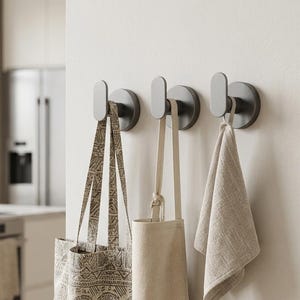 May include: Three matte dark grey wall-mounted hooks with a circular base and an oval-shaped front hook. A patterned tote bag, a plain beige apron, and a textured light beige cloth are hanging from the hooks on a smooth off-white wall.