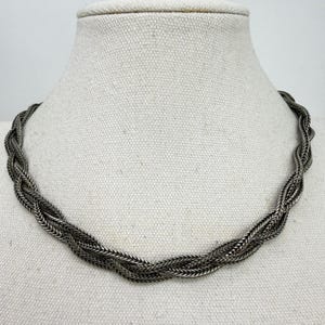 May include: A silver-toned necklace featuring a multi-strand braided design. The chain is composed of several interwoven metallic links that create a textured, rope-like appearance. The necklace rests in a circular shape with a consistent finish.