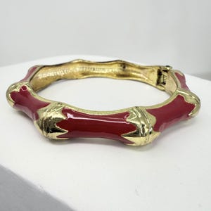Op de afbeelding: Een goudkleurige scharnierende armband met een bamboe-ontwerp. De armband heeft segmenten van glanzend rood email en goudkleurig metaal. De armband is een opvallend sieraad.
