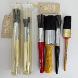 Può includere: Una collezione di sei pennelli per stencil con setole scure e vari manici. Due pennelli sono all'interno di tubi di plastica trasparente con etichette Country Colors Stencil Brush. Gli altri quattro hanno manici rossi, gialli, neri e in legno.