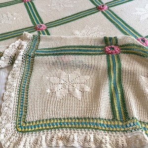 Pode incluir: Uma manta de crochê na cor creme com um padrão de grade definido por listras verdes, amarelas e azuis. As interseções são acentuadas com pequenos motivos florais rosa em relevo, e o centro de cada quadrado exibe um design em forma de estrela.