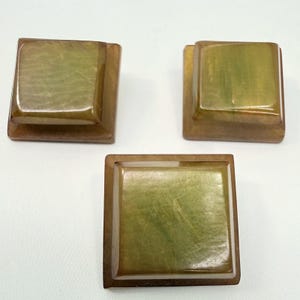 May include: Three square, decorative cabinet knobs. The knobs have a light green center and a brown frame. The knobs are made of a glossy material and are arranged on a white surface.
