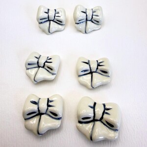 May include: Six ceramic buttons shaped like bows, arranged in three rows of two. Each button is off-white with dark blue hand-painted lines accentuating the folds and center knot of the bow design. The surface of each button is smooth and glossy.