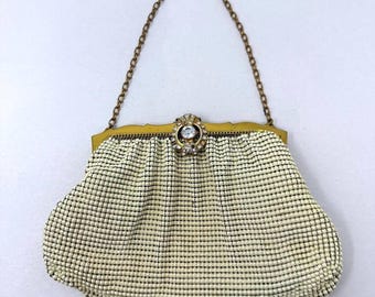 May include: A cream-colored mesh handbag featuring a gold-tone metal frame and a matching chain strap. The center of the frame is adorned with a circular decorative clasp set with clear rhinestones, providing a secure closure for the pouch.