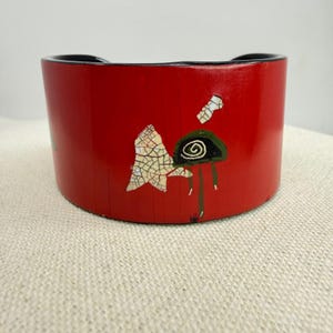 May include: A red, cylindrical wooden container with a black rim. The container is decorated with a white, cracked leaf design and a green and black spiral design with green lines. The container sits on a beige surface.