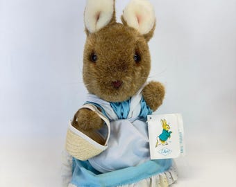 May include: A brown plush rabbit toy with long ears, wearing a light blue dress with a white apron and a white eyelet lace trim at the hem. The rabbit holds a small woven basket and has a white tag attached that features a graphic of a rabbit.