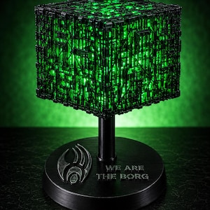 May include: A black cube-shaped lamp with intricate mechanical surface details illuminated by a bright green light. The cube is mounted on a black cylindrical stand with a circular base featuring a metallic emblem and the text WE ARE THE BORG.