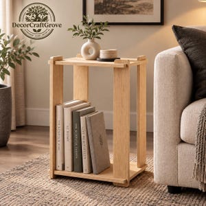 May include: A light-toned wooden side table with two tiers. The top surface holds a white ceramic vase with greenery and a small round container. The bottom shelf stores four books titled The Kinfolk Home, At Home, The Kinfolk Garden, and Live Beautifully.