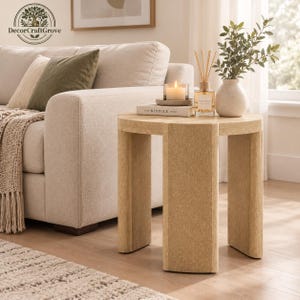 May include: A round, light-tan side table with three thick, curved legs. On the table sits a white candle in a glass holder, a book titled The Kinfolk Home, a glass reed diffuser with wooden sticks, and a small white vase with green leaves.